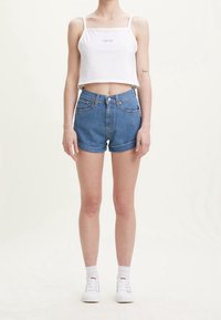 Persona vistiendo un top corto blanco, pantalones cortos de denim azul enrollados, calcetas blancas y zapatillas blancas, de pie contra un fondo liso.