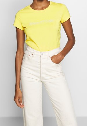 Gul bomuld T-shirt med rund hals og korte ærmer. Har diskret hvidt logotekst på fronten. Kombineret med højtaljede hvide denimjeans.