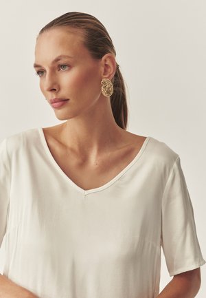 Biely top s krátkym rukávom a V-výstrihom. Nosí sa s veľkými oválnymi náušnicami so zlatou textúrou. Neutrálne pozadie, hladká textúra látky.