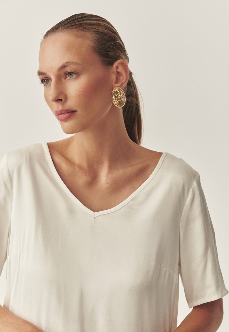 Valge lühikeste varrukatega särk V-kaeluse disainiga. Kanda koos suurte kuldsete tekstuuriga ovaalsete kõrvarõngastega. Neutraalne taust, sile kangatekstuur.