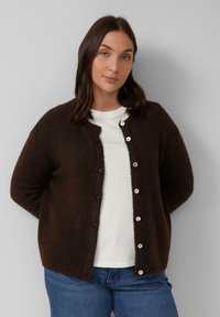 Braune Strickjacke mit rundem Ausschnitt, strukturierter Stoff und silbernen Knöpfen vorne. Getragen über einem weißen Hemd.