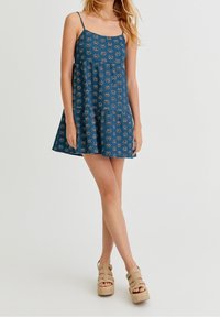 Nuori nainen, joka pukeutuu siniseen kukalliseen hihattomaan mekkoon ja vaaleisiin platform-sandaaleihin, seisoo jalat ristissä valkoista taustaa vasten.