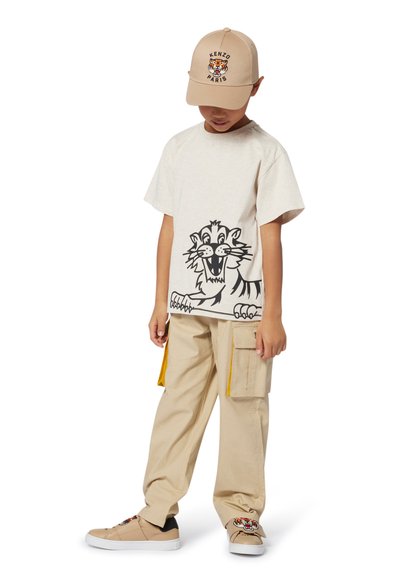 T-shirt en coton beige avec un graphisme de tigre cartoon noir, associé à un pantalon cargo beige avec des accents jaunes et des baskets beiges avec un détail de tigre.