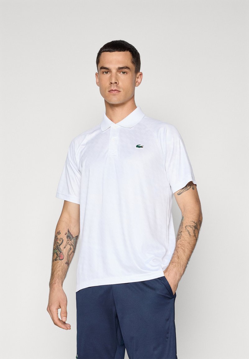 Polo shirt blanc avec un col classique, des manches courtes et un petit logo vert. Le tissu semble lisse avec un motif subtil.