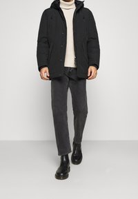 Giacca pantalone nera con cappuccio, zip frontali e bottoni a pressione. Indossata sopra un dolcevita beige e jeans grigio scuro. Stivaletti neri.