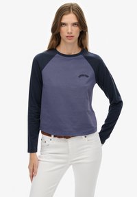 Młoda kobieta z długimi włosami, ubrana w dwukolorową bluzkę z długim rękawem w odcieniach granatu i fioletu oraz białe spodnie z brązowym plecionym paskiem.