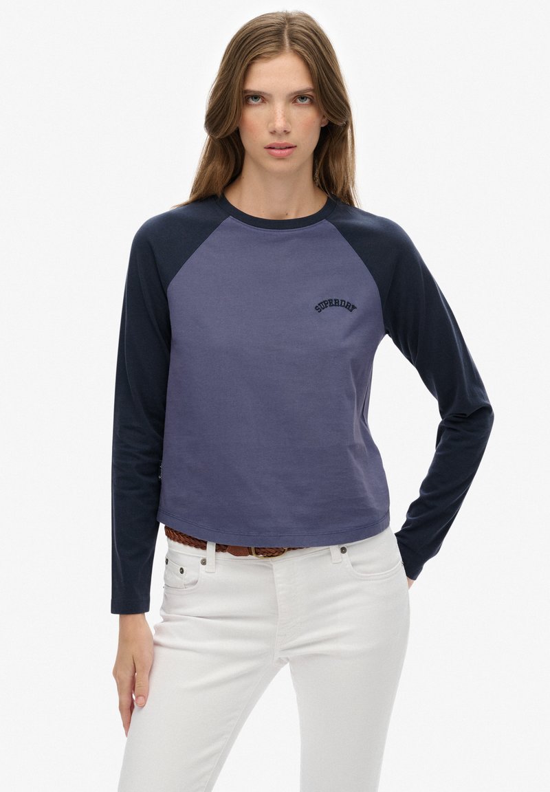 Młoda kobieta z długimi włosami, ubrana w dwukolorową bluzkę z długim rękawem w odcieniach granatu i fioletu oraz białe spodnie z brązowym plecionym paskiem.