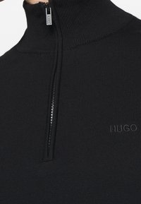 Suéter negro de punto acanalado con media cremallera y cuello alzado, que presenta el sutil bordado "HUGO" en el pecho.