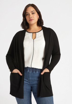 Cardigan noir avec des poignets côtelés et des poches, porté sur un haut léger avec un décolleté en keyhole. Style décontracté, tissu texturé, coupe relax.