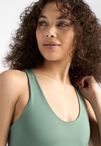 Bañador verde, de corte bajo, con una textura suave y tirantes anchos, que acentúan el escote y el área de los hombros. Se adivinan unos rizos en el cabello.