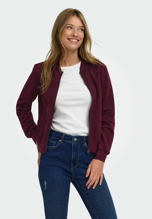 Lächelnde Frau, die eine weinrote Jacke mit Reißverschluss über einem weißen Hemd und blauen Jeans trägt und mit einer Hand in der Hüfte steht.