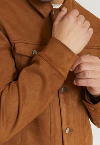 Chaqueta de ante marrón con cierres a presión, un collar puntiagudo y bolsillos en el pecho. La textura es suave y lisa, con un diseño ajustado.