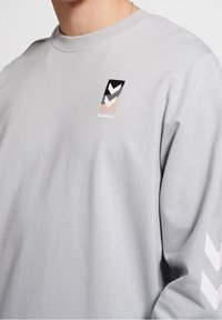 Felpa grigia chiaro con un piccolo logo verticale nero, bianco e beige e chevron sul petto e chevron bianchi sulla manica.