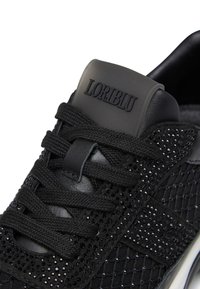 Black sneaker featuring a textured mesh upper with studded accents, lace closure, and a rubberized logo patch on the tongue.