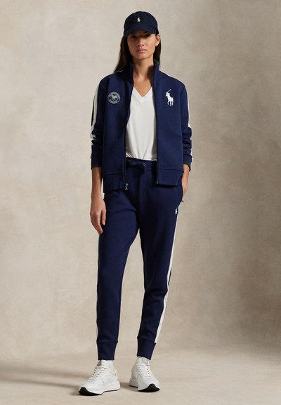 Marinblå träningsjacka med vita ränder och en logotyp, matchad med byxor och keps i samma stil. Outfitten har dragkedja och elastiska mudd.