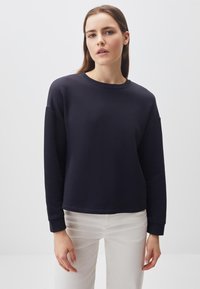 Felpa blu navy con girocollo, spalle basse, maniche lunghe e polsini a costine. Tessuto liscio, abbinata a pantaloni chiari.