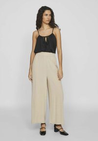 Top negro sin mangas con un detalle de lazo y pantalones anchos beige. El conjunto presenta un ajuste relajado y una tela ligera. Zapatos de tacón negros.