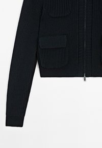 Cardigan nero lavorato a maglia con maniche lunghe, cerniera frontale e tasca con pattina sul lato sinistro su sfondo bianco.