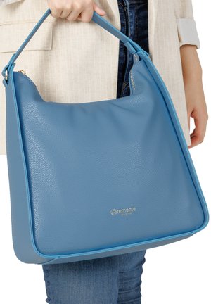 Personne tenant un grand sac à main en cuir bleu avec une seule bandoulière, portant une veste beige et un jean bleu.