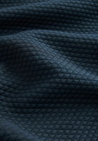 Tissu texturé bleu foncé avec un motif matelassé en losange créant des plis subtils et des ombres à sa surface.