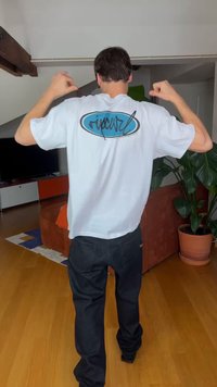 Homme de dos, portant un t-shirt blanc avec un logo bleu et noir, pantalon noir, debout dans un salon avec un sol en bois et une plante en pot.