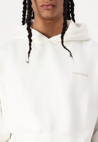 Sudadera blanca con capucha de textura suave, que cuenta con un bolsillo tipo canguro y un discreto logo beige en el pecho. Los cordones ajustables completan el diseño.