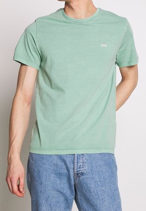 Homme portant un t-shirt vert clair uni à col rond et manches courtes, ainsi qu'un jean en denim bleu, sur fond blanc.