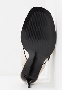 Zapato de tacón alto beige con suela negra, con punta afilada y tacón de aguja delgado. Superficie texturizada y diseño minimalista.