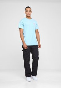 T-shirt en coton bleu clair à manches courtes, présentant un petit logo rose sur la poitrine, associé à un pantalon cargo noir et des baskets blanches.