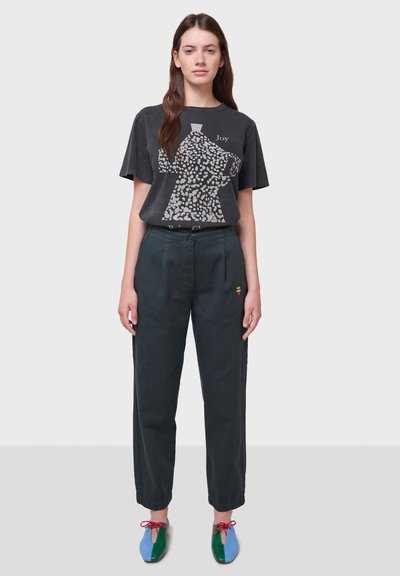 T-shirt grafico nero con design leopardato, abbinato a pantaloni su misura verde scuro con un piccolo logo colorato. Scarpe colorate e strutturate.