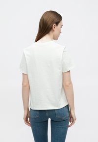 Biele bavlnené tričko s krátkym rukávom, priamym strihom a okrúhlym výstrihom, zobrazené zozadu, v kombinácii s modrými rifľami.