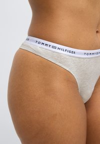 Μπεζ βαμβακερό στρινγκ εσώρουχο με λευκή ελαστική ζώνη μέσης που φέρει μαύρη επιγραφή "TOMMY HILFIGER" κατά μήκος της κορυφής. Λείος υφής.