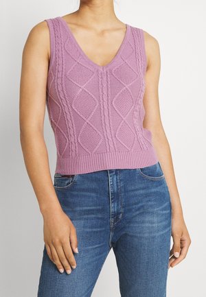 Person iført ærmeløs lavendel strikket sweatervest med diamantmønstre og blå jeans, stående mod en ensfarvet lys baggrund.