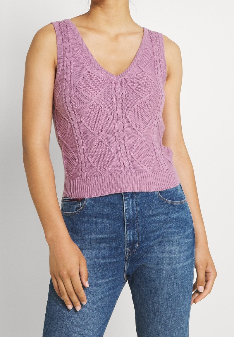 Personne portant un gilet en tricot lavande sans manches avec des motifs en losange et un jean bleu, debout devant un fond clair uni.