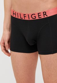 Crne pamučne bokserice s širokim ružičastim pojasom na kojem je crnim slovima ispisano "HILFIGER". Glatka tekstura, prilagođeni dizajn i dužina do sredine bedara.