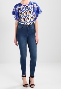 Blå mønstret top med flagrende ærmer, geometrisk design i gul og sort. Tætsiddende mørke jeans, højhælede sorte sandaler.