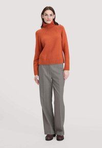 Frau trägt einen orangefarbenen Rollkragenpullover, graue weite Hosen und dunkelbraune Schuhe, während sie vor einem schlichten hellen Hintergrund steht.