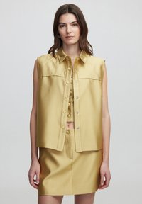 Sleeveless, light yellow shirt with a pointed collar and snap buttons, paired with a coordinating mini skirt in the same fabric and color.