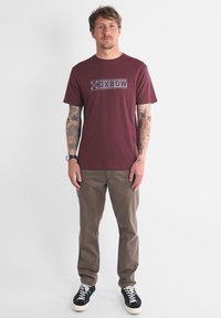 T-shirt à col rond bordeaux avec le logo "OXBOW", associé à un pantalon kaki et des baskets noires avec des lacets blancs, se tenant sur un fond uni.