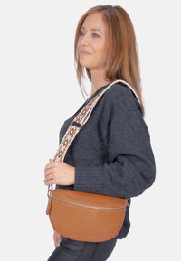 Braune Leder-Crossbody-Tasche mit runder Form, ausgestattet mit einer strukturierten Oberfläche und einem gemusterten Gurt in neutralen Farbtönen.