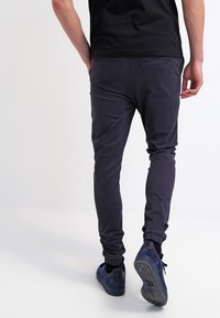 Osoba v námořnických jogger kalhotách a modrých teniskách, kráčející pryč na bílém pozadí.