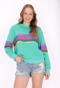 Türkiser Pullover mit kräftigen horizontalen Streifen in Pink, Blau und Gelb. Lange Ärmel und lockere Passform. Kombiniert mit abgetragenen Denim-Shorts.