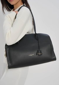 Femme en pull blanc portant un grand sac  à main en cuir noir Lancaster Paris sur l'épaule, sur fond uni.