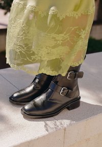 Stivali neri in pelle fino alla caviglia con due fibbie in argento, punta arrotondata e tacco a blocco basso, posizionati su una superficie di pietra testurizzata.