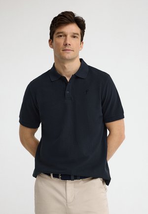 Mand står med hænderne bag ryggen iført sort kortærmet poloshirt og beige bukser mod en ensfarvet lys baggrund.