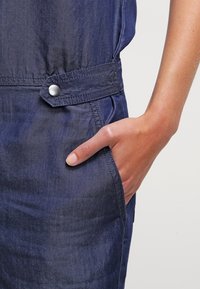 Main reposant dans la poche avant d'un vêtement en denim bleu foncé avec un détail de bouton-pression argenté à la ceinture.