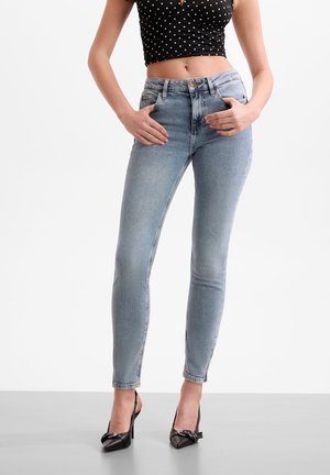 Femme portant un jean skinny bleu clair, des talons hauts noirs pointus et un crop top noir à pois blancs sur fond blanc.
