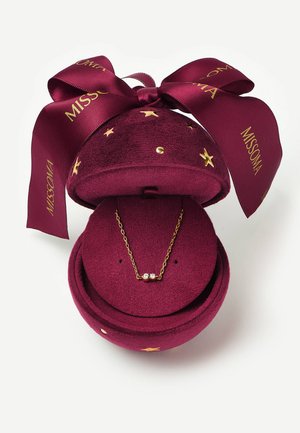 Boîte à bijoux ronde en velours bordeaux avec des étoiles dorées et un ruban, contenant un délicat collier en chaîne dorée avec trois petites pierres précieuses.