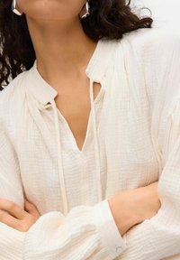 Vit texturerad blus med V-halsringning, dragsko-detalj och voluminösa ärmar. Har knoppar vid manschetterna och subtila veck.