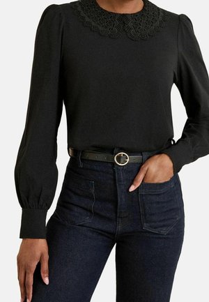 Blouse noire à manches longues avec un col en dentelle, associée à un jean foncé taille haute et une ceinture noire avec une boucle ronde dorée.